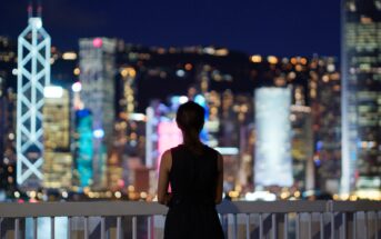 A person stands on a balcony at night, overlooking a city skyline with brightly lit skyscrapers. The city lights are blurred, creating a colorful bokeh effect, while the person is silhouetted against the vibrant background.