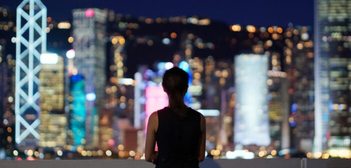 A person stands on a balcony at night, overlooking a city skyline with brightly lit skyscrapers. The city lights are blurred, creating a colorful bokeh effect, while the person is silhouetted against the vibrant background.