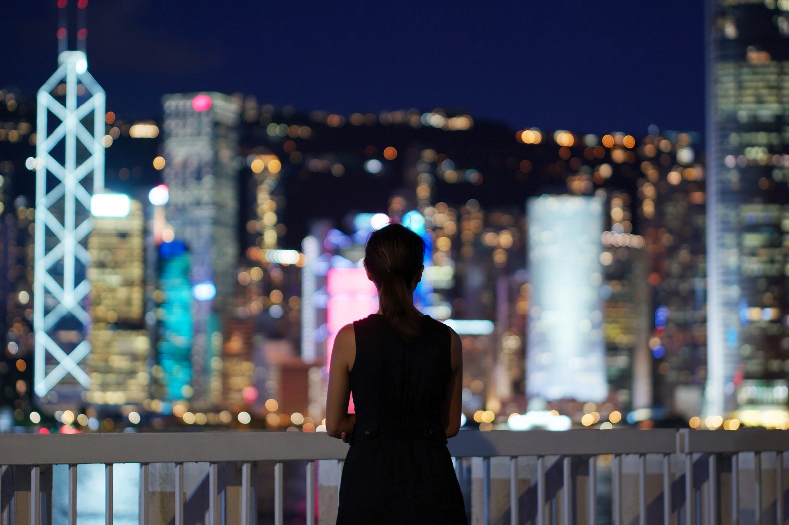 A person stands on a balcony at night, overlooking a city skyline with brightly lit skyscrapers. The city lights are blurred, creating a colorful bokeh effect, while the person is silhouetted against the vibrant background.