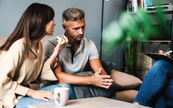 A couple sits on a couch, engaged in conversation with an unseen therapist. The woman gestures as she speaks, while the man listens attentively. A coffee cup is on the table. The setting is a cozy, modern therapy room with books and plants in the background.