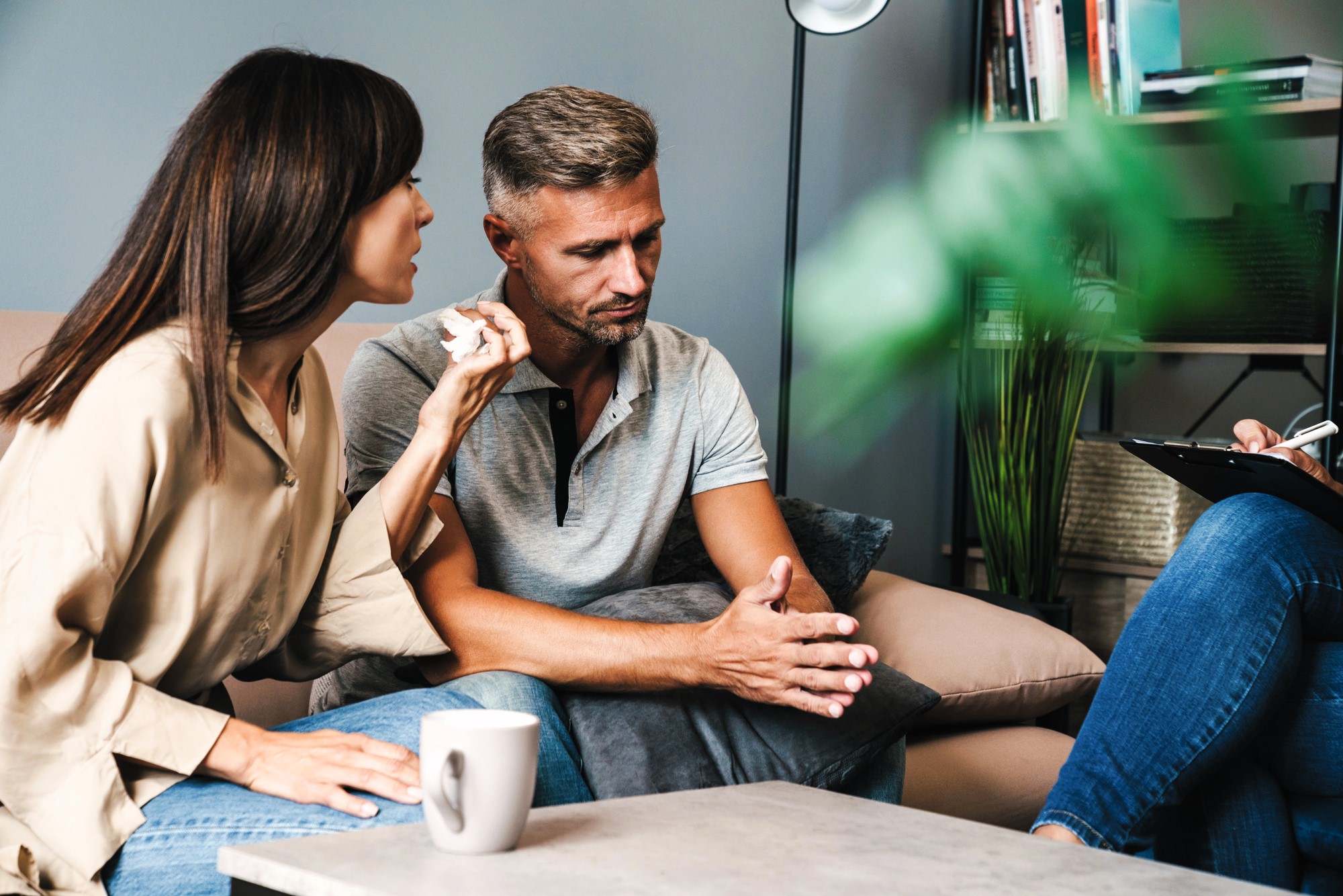 A couple sits on a couch, engaged in conversation with an unseen therapist. The woman gestures as she speaks, while the man listens attentively. A coffee cup is on the table. The setting is a cozy, modern therapy room with books and plants in the background.