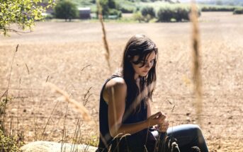 A woman with long hair, wearing a dark tank top and jeans, sits on a rock in a field. She is looking down at her hands. The background shows a wide, sunlit landscape with trees and a clear sky.