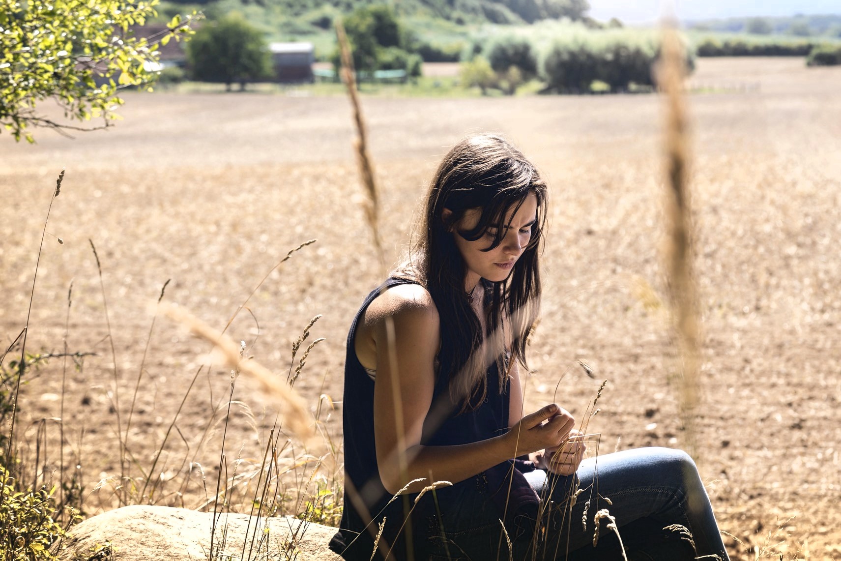 A woman with long hair, wearing a dark tank top and jeans, sits on a rock in a field. She is looking down at her hands. The background shows a wide, sunlit landscape with trees and a clear sky.