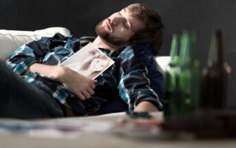 A man with a beard, wearing a plaid shirt, sleeps on a couch holding a photo of a woman. Several empty green bottles are in the foreground, and a remote control lies beside him. The background is dark, contrasting with the scene.