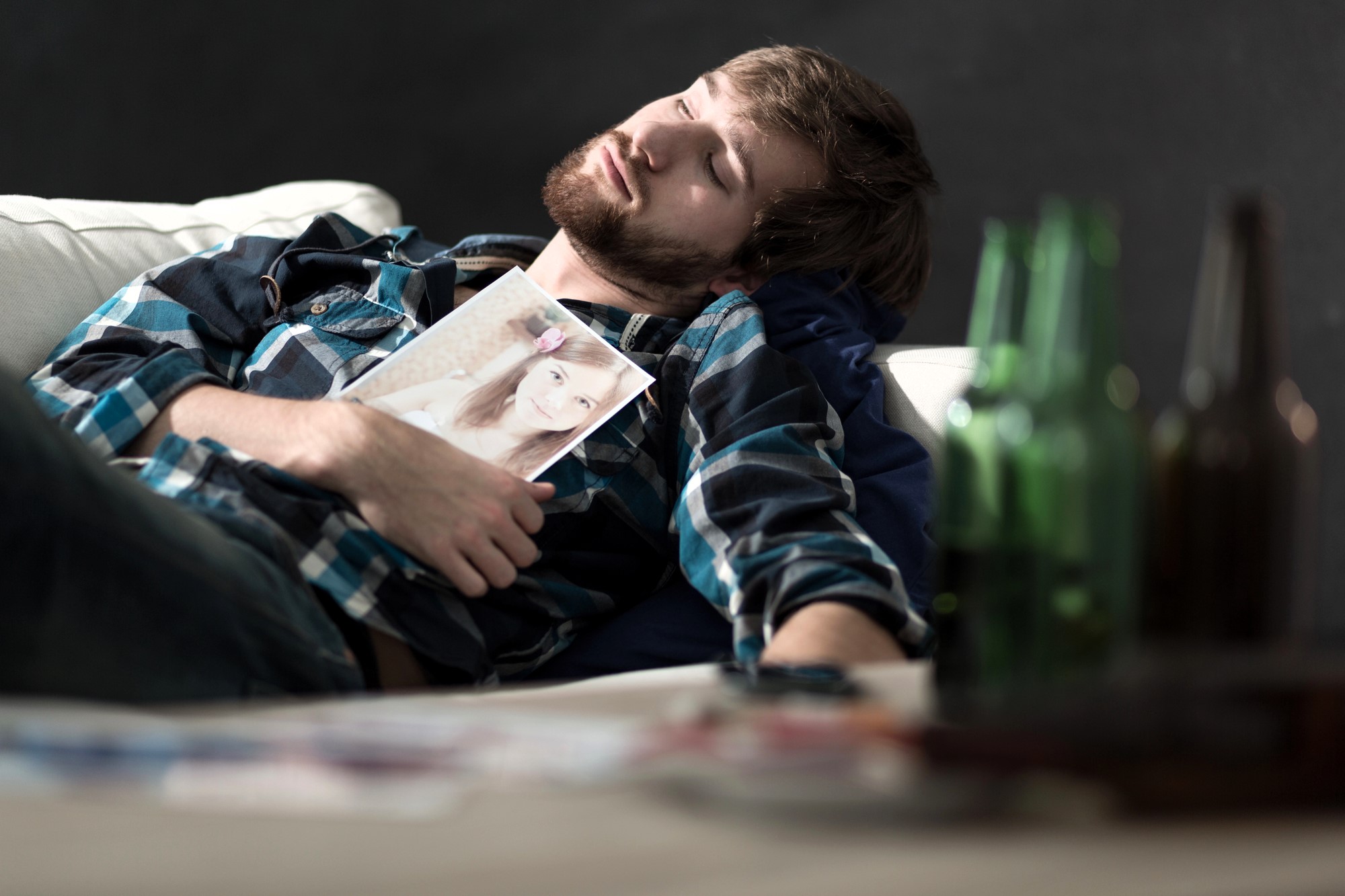 A man with a beard, wearing a plaid shirt, sleeps on a couch holding a photo of a woman. Several empty green bottles are in the foreground, and a remote control lies beside him. The background is dark, contrasting with the scene.