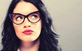 Woman with long black hair wearing cat-eye glasses and red lipstick, looking upwards thoughtfully against a neutral background.