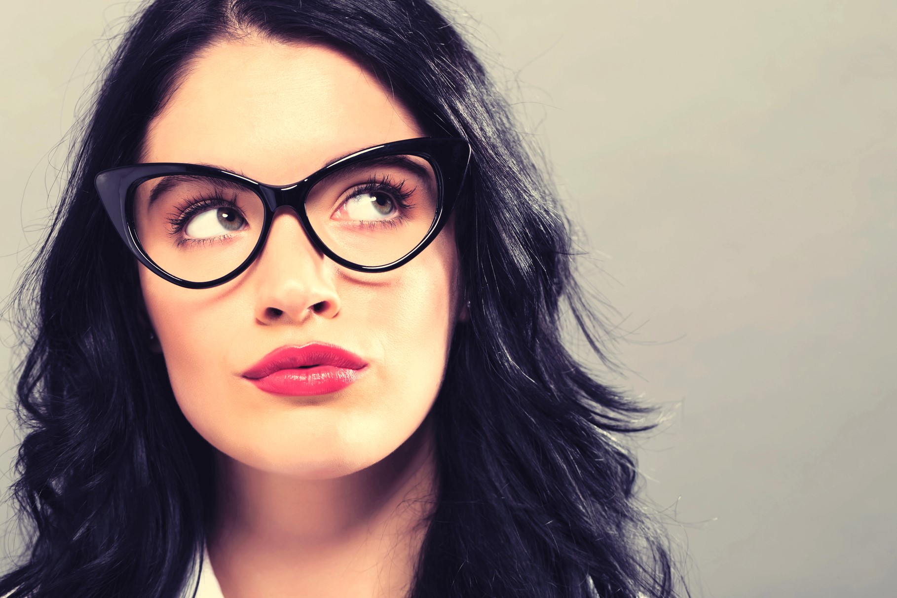 Woman with long black hair wearing cat-eye glasses and red lipstick, looking upwards thoughtfully against a neutral background.