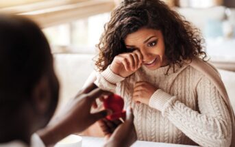 A woman with curly hair and a beige sweater appears tearful and surprised as she looks at a person holding an open ring box in front of her at a restaurant table. The scene suggests a romantic proposal.