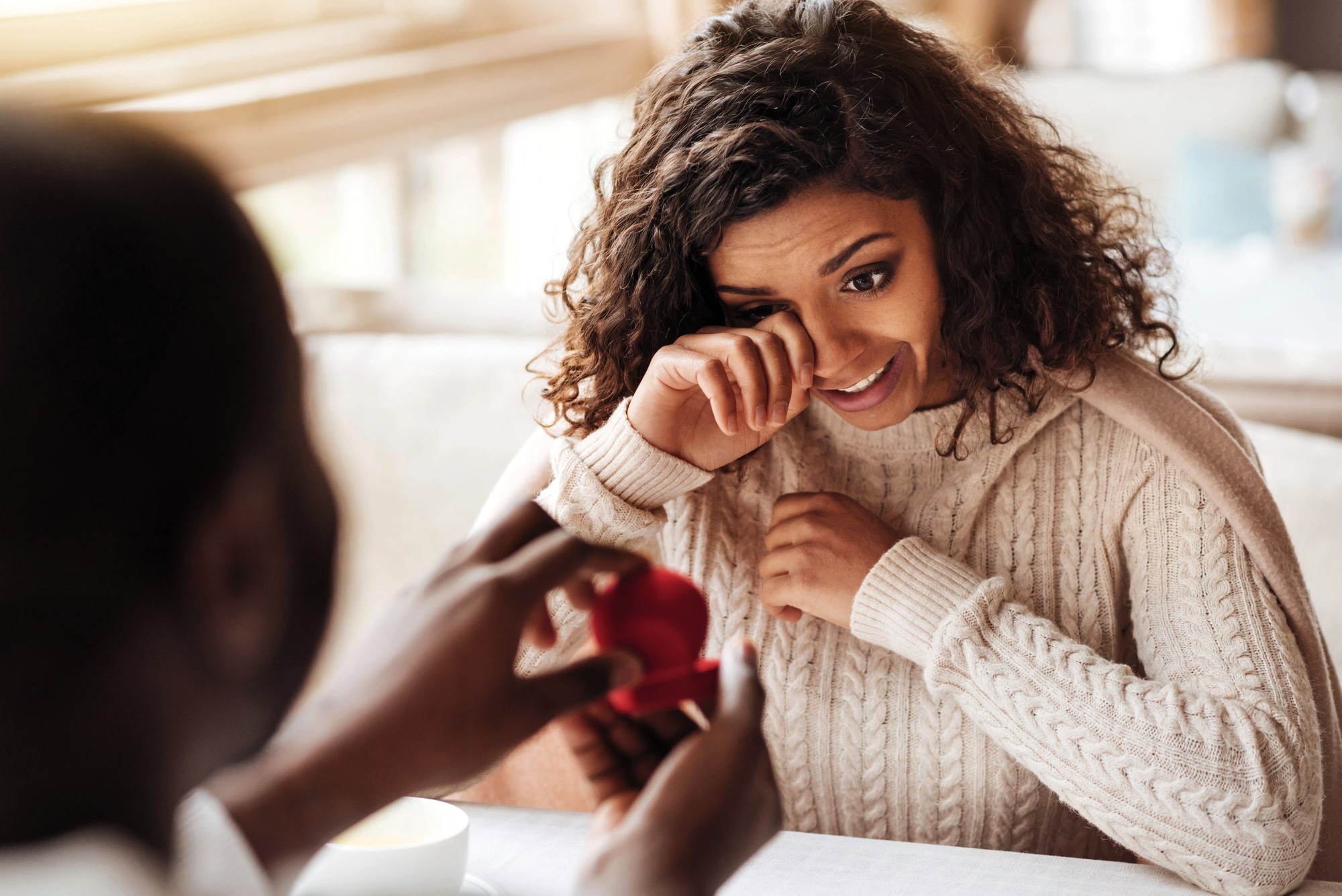 A woman with curly hair and a beige sweater appears tearful and surprised as she looks at a person holding an open ring box in front of her at a restaurant table. The scene suggests a romantic proposal.