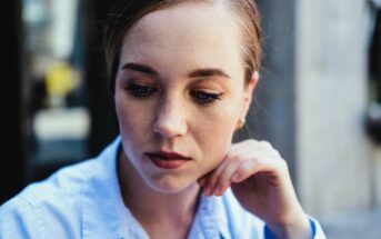 A woman with light brown hair pulled back is gazing downward, resting her chin on her hand. She is wearing a light blue collared shirt and appears to be in deep thought or contemplation, with a blurred background behind her.