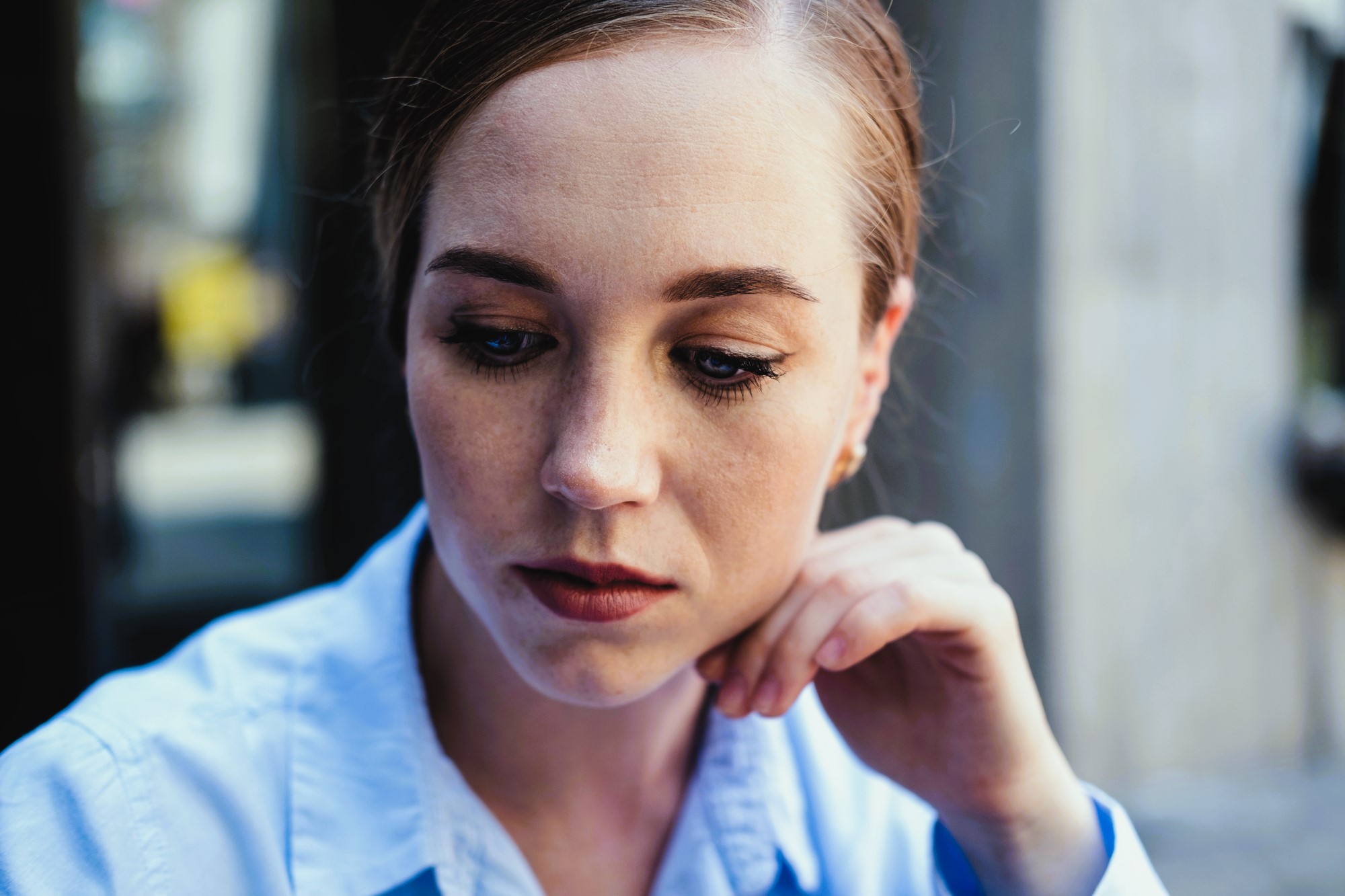 A woman with light brown hair pulled back is gazing downward, resting her chin on her hand. She is wearing a light blue collared shirt and appears to be in deep thought or contemplation, with a blurred background behind her.