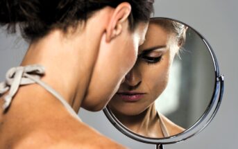 A woman with brown hair tied back, wearing a light-colored tank top, is looking down into a round mirror. Her reflection shows a thoughtful expression. The background is neutral, creating a serene atmosphere.