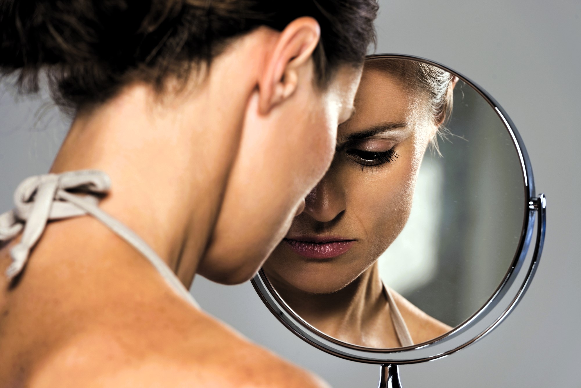 A woman with brown hair tied back, wearing a light-colored tank top, is looking down into a round mirror. Her reflection shows a thoughtful expression. The background is neutral, creating a serene atmosphere.