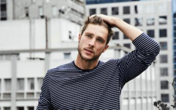 A man with short brown hair and a beard is wearing a black and white striped shirt. He is standing on a rooftop, one hand on his head, with a backdrop of urban buildings.