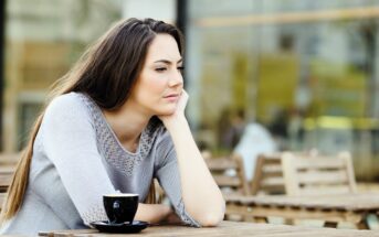 A woman with long brown hair sits at an outdoor cafe, resting her chin on her hand, looking thoughtful. She is wearing a grey sweater and there is a cup of coffee on the wooden table in front of her. Blurred background of the cafe setting.