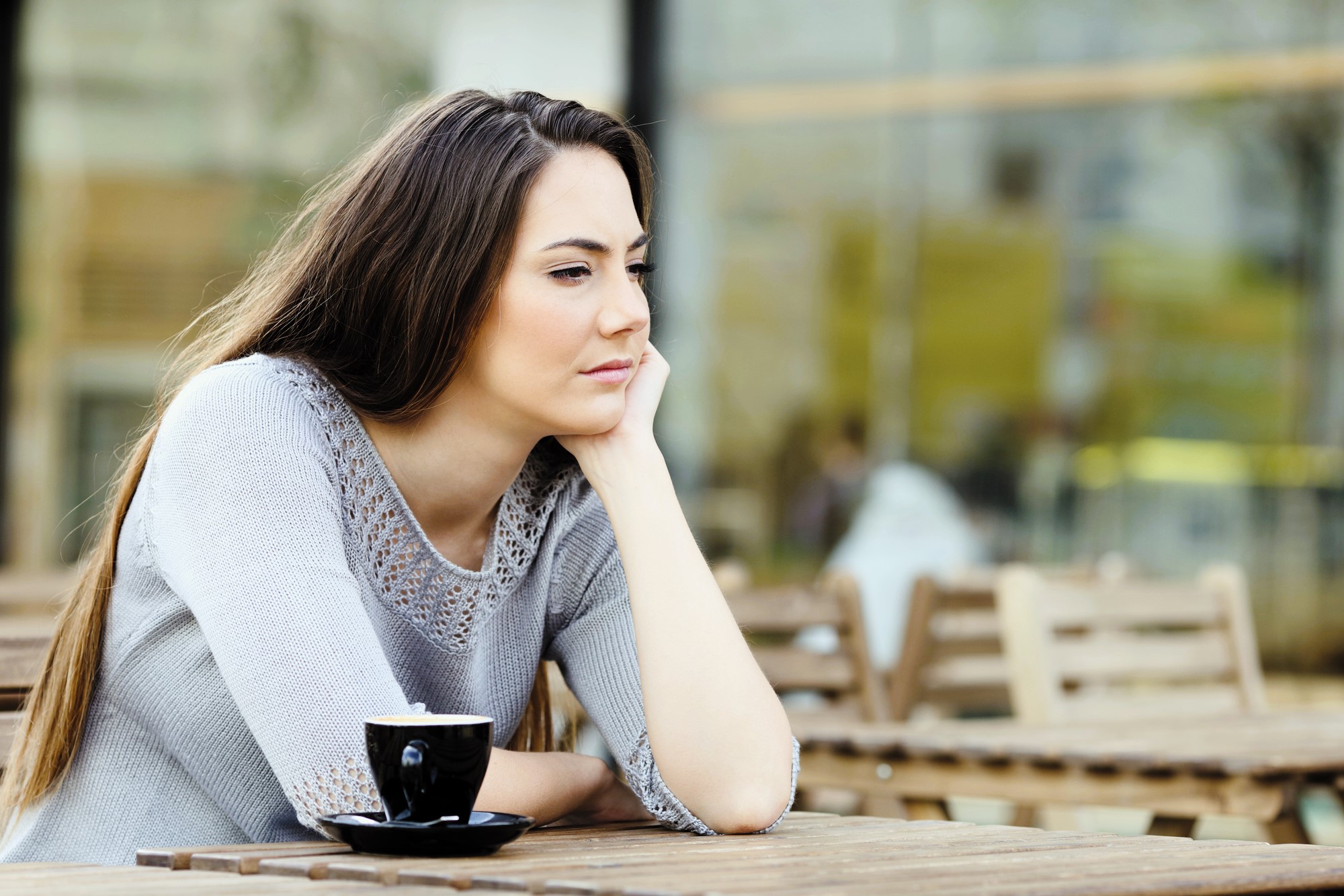 A woman with long brown hair sits at an outdoor cafe, resting her chin on her hand, looking thoughtful. She is wearing a grey sweater and there is a cup of coffee on the wooden table in front of her. Blurred background of the cafe setting.