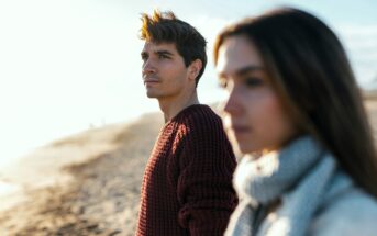 A man in a red sweater and a woman in a gray scarf stand on a sandy beach, gazing thoughtfully into the distance. The sunlight creates a warm glow, and the ocean is visible in the background.