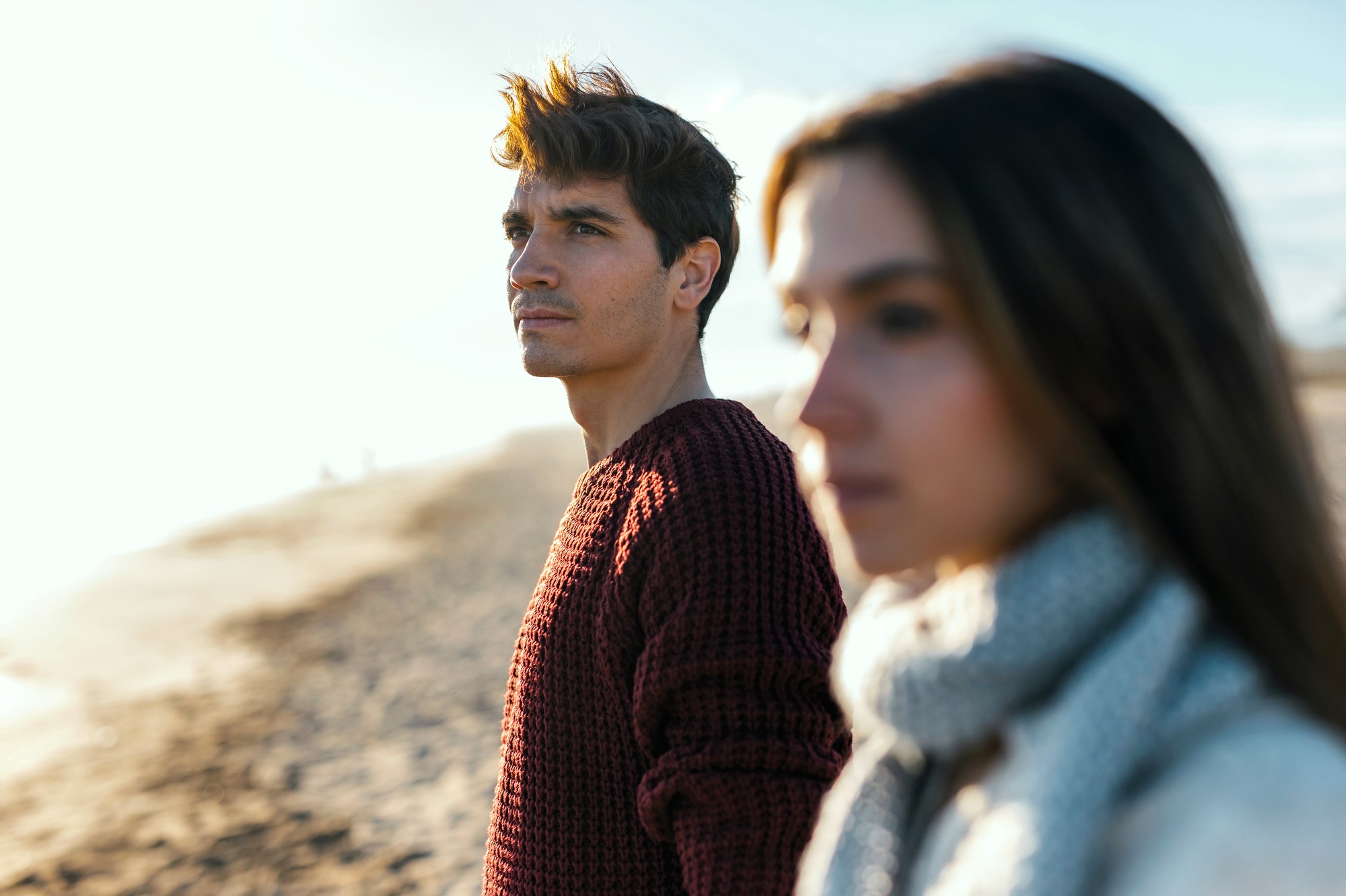 A man in a red sweater and a woman in a gray scarf stand on a sandy beach, gazing thoughtfully into the distance. The sunlight creates a warm glow, and the ocean is visible in the background.