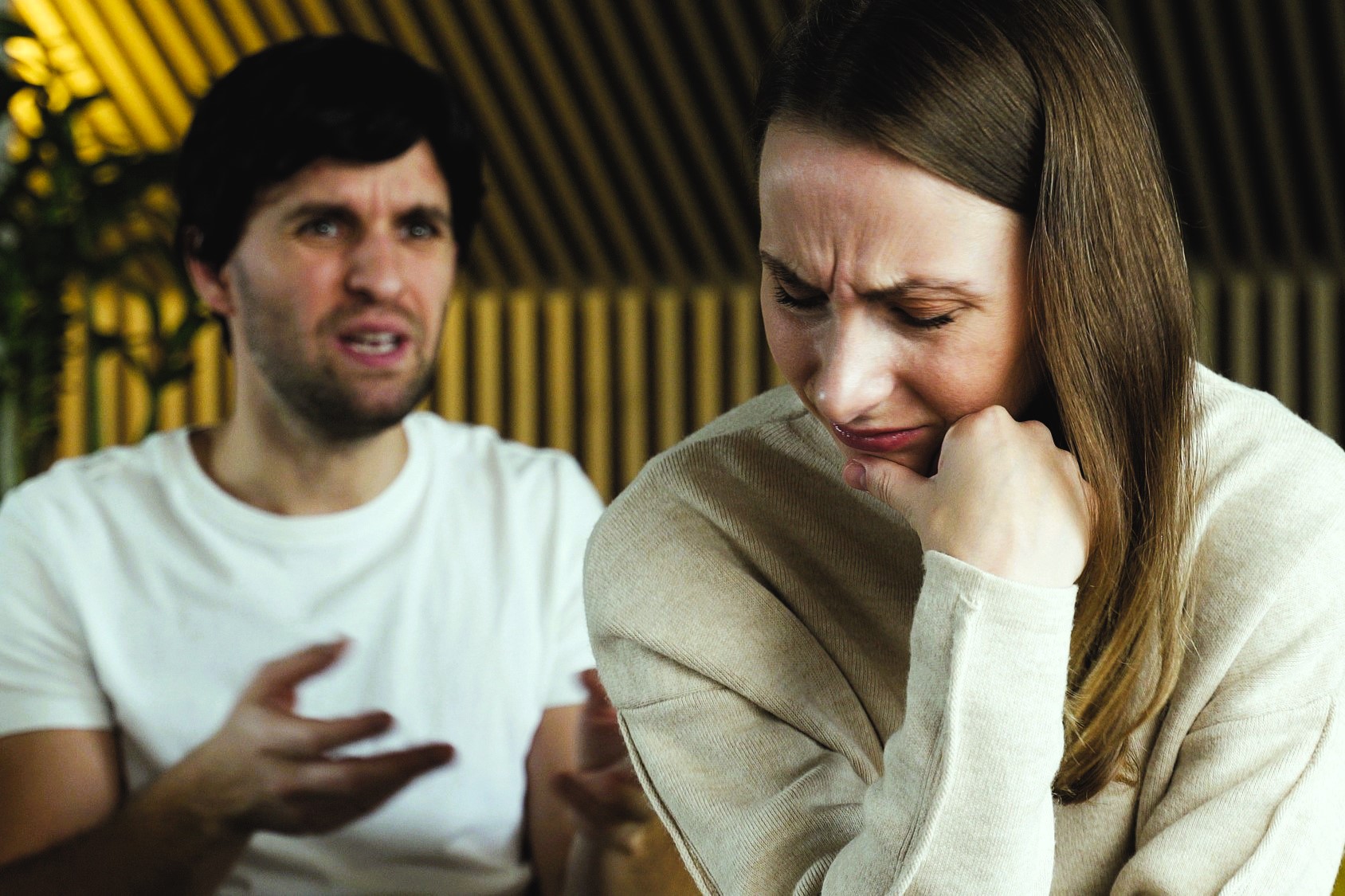 A woman in the foreground looks distressed, resting her chin on her hand. A man in the background appears to be speaking animatedly. They are indoors, with a dimly lit background.