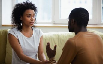 A woman with curly hair and a gray shirt sits on a couch, holding hands and having a conversation with a man wearing glasses and a brown sweater. They are indoors, near a large window.