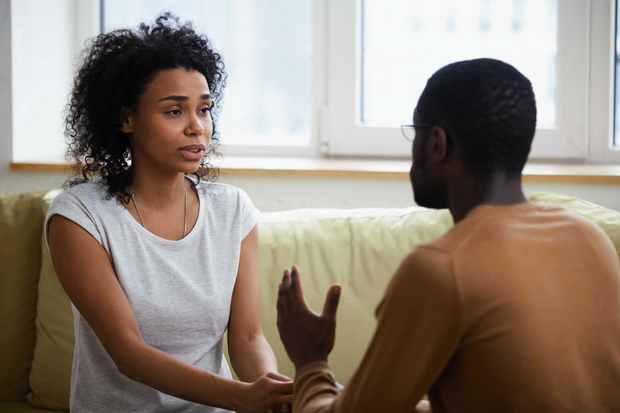 A woman with curly hair and a gray shirt sits on a couch, holding hands and having a conversation with a man wearing glasses and a brown sweater. They are indoors, near a large window.