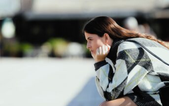 A woman with long brown hair sits in profile, resting her chin on her hand. She is wearing a patterned sweater and appears to be deep in thought, with a blurred outdoor background.