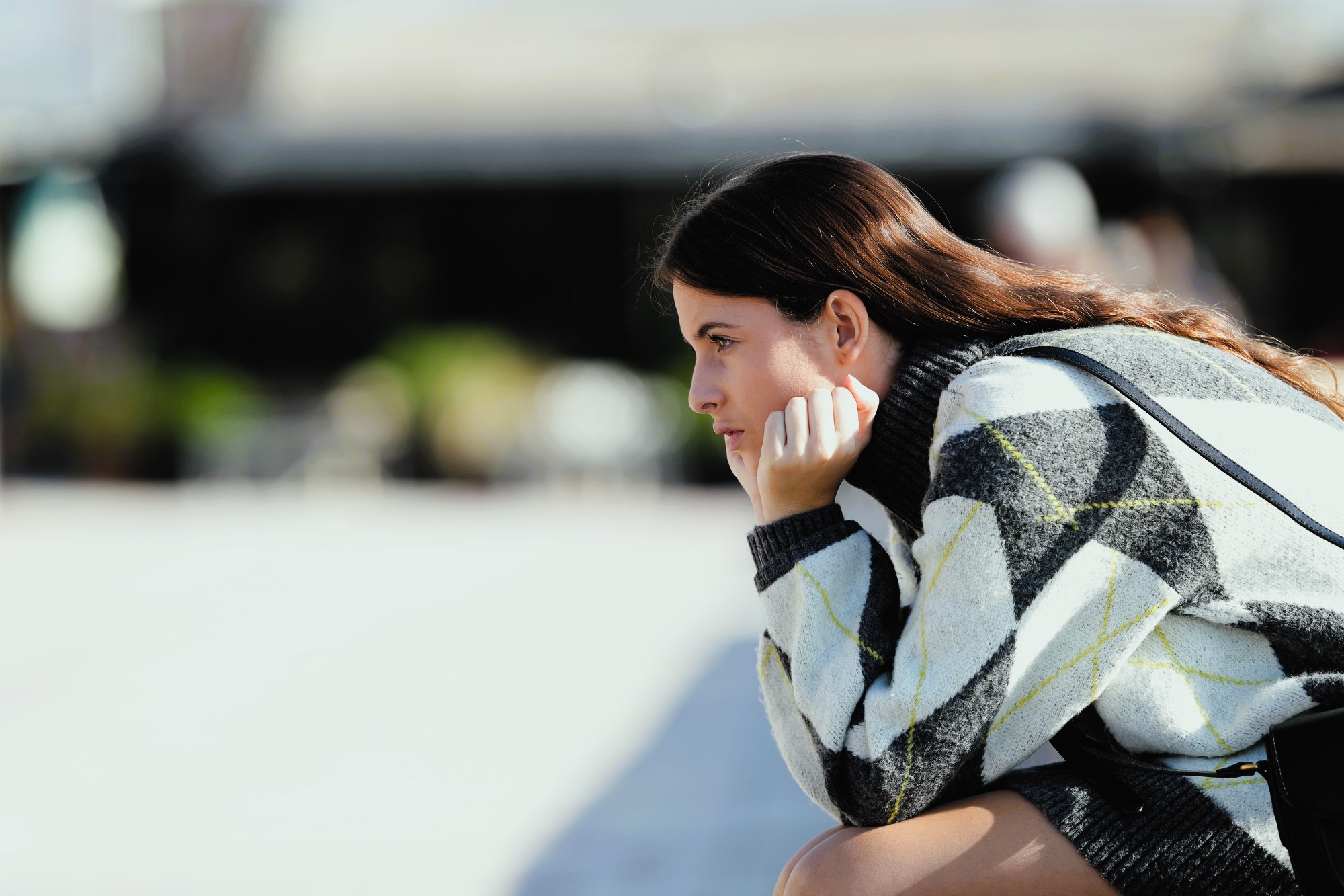 A woman with long brown hair sits in profile, resting her chin on her hand. She is wearing a patterned sweater and appears to be deep in thought, with a blurred outdoor background.