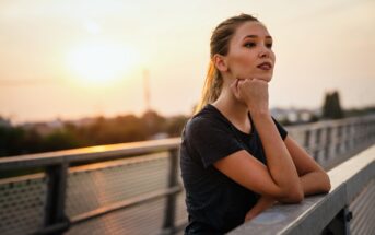 A woman with long hair tied back is leaning on a railing, gazing thoughtfully into the distance. She is wearing a dark shirt. The background shows a soft sunset over a blurred cityscape.