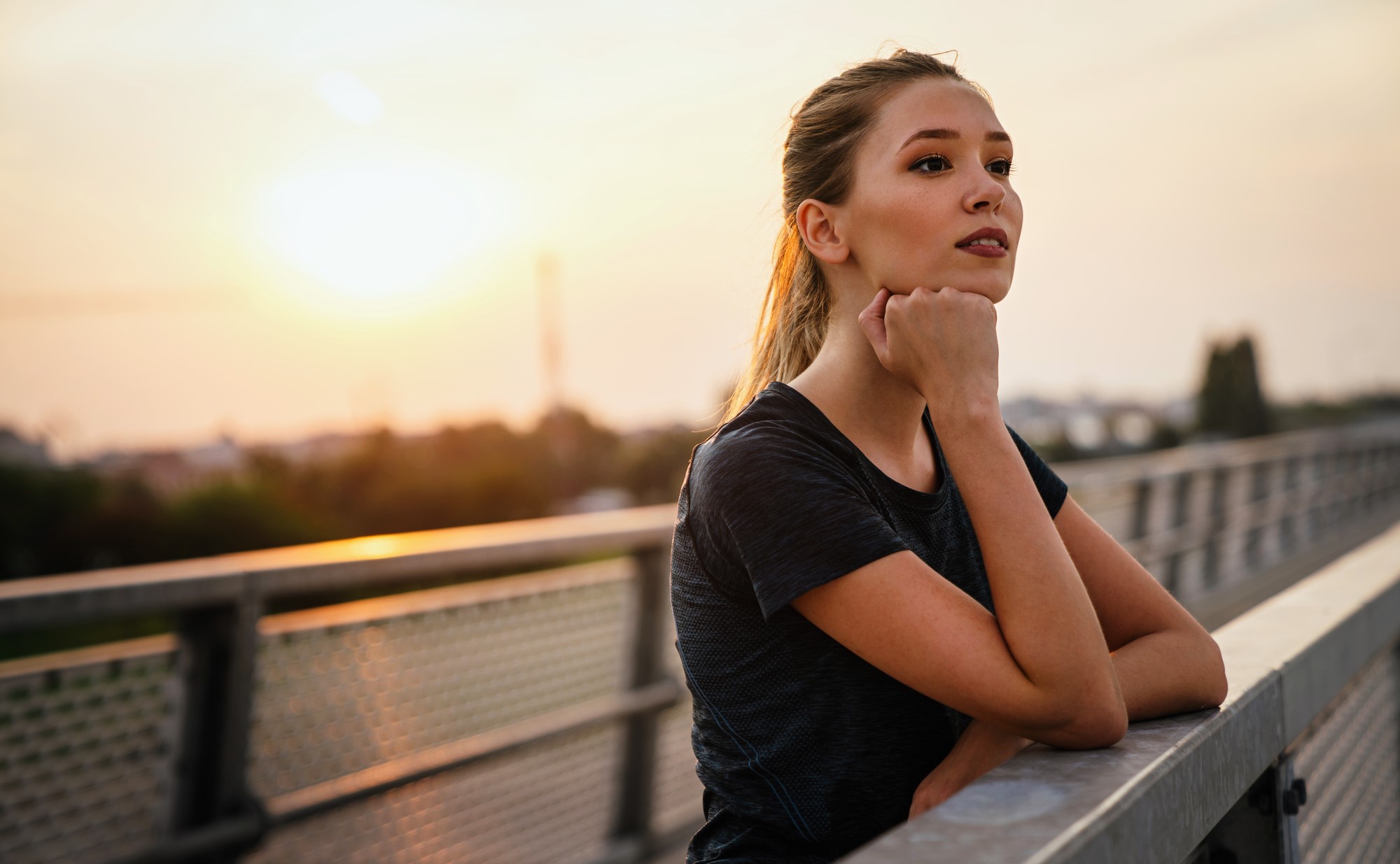 A woman with long hair tied back is leaning on a railing, gazing thoughtfully into the distance. She is wearing a dark shirt. The background shows a soft sunset over a blurred cityscape.