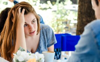 A woman with long brown hair, wearing a gray shirt, sits at a table, resting her head on her hand. She looks towards a person across from her. In front of her is a cup of coffee and a floral centerpiece. The background is blurred.