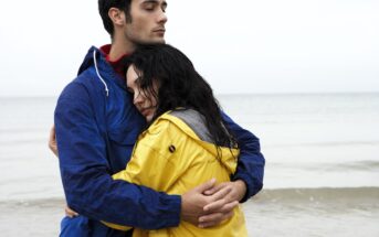 A couple stands on a beach in overcast weather. The man wears a blue jacket, holding the woman close to him. She wears a yellow jacket and rests her head on his chest, eyes closed. The ocean is calm in the background.