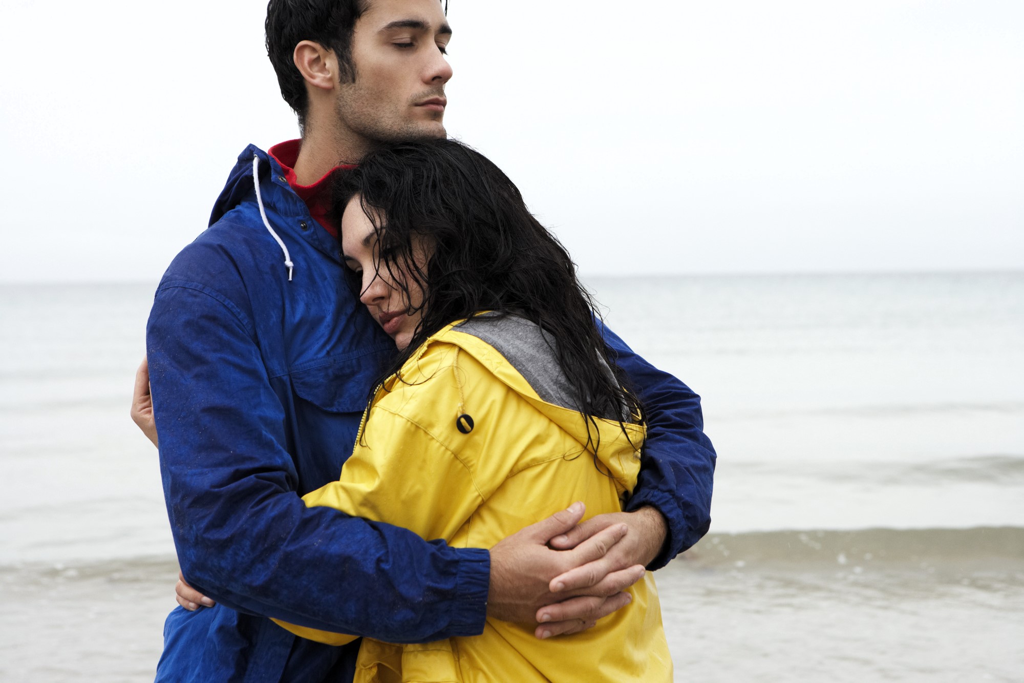 A couple stands on a beach in overcast weather. The man wears a blue jacket, holding the woman close to him. She wears a yellow jacket and rests her head on his chest, eyes closed. The ocean is calm in the background.