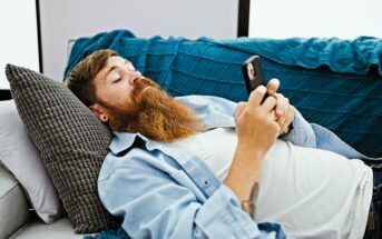 A man with a long beard, lying on a sofa, looks at his smartphone. He is resting his head on a gray pillow and wearing a light blue shirt over a white t-shirt. A blue quilt covers part of the sofa.