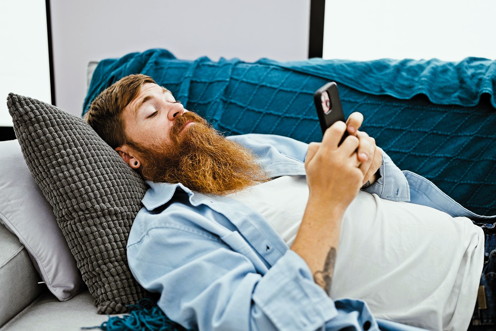 A man with a long beard, lying on a sofa, looks at his smartphone. He is resting his head on a gray pillow and wearing a light blue shirt over a white t-shirt. A blue quilt covers part of the sofa.