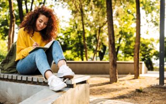 A person with curly red hair, wearing a yellow sweater and jeans, sits on a bench outdoors, writing in a notebook. The setting is a sunlit park with trees and a paved path in the background.