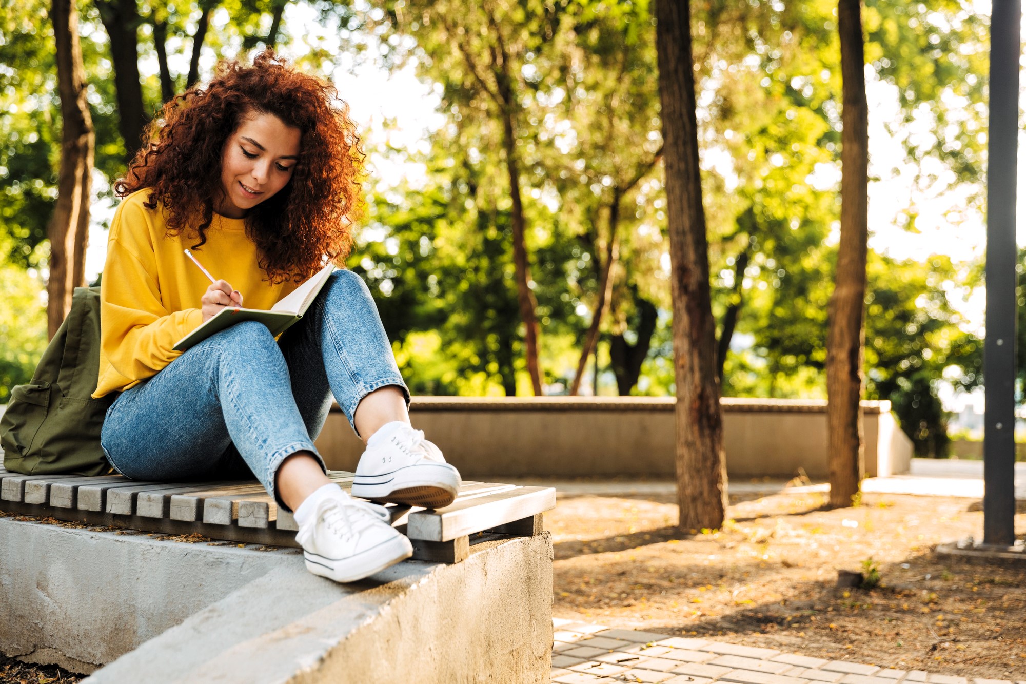 A person with curly red hair, wearing a yellow sweater and jeans, sits on a bench outdoors, writing in a notebook. The setting is a sunlit park with trees and a paved path in the background.