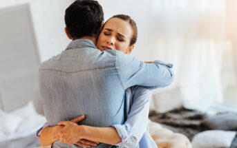 A woman and a man embrace in a comforting hug. The woman closes her eyes, appearing relieved or emotional. They stand in a softly lit room with a blurred background that includes a bed and a cozy ambiance.