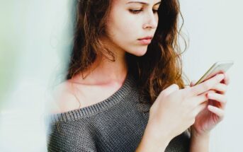 A woman with long brown hair and an off-the-shoulder gray sweater is intently using a smartphone. She is positioned against a blurred white background, focusing on the screen.