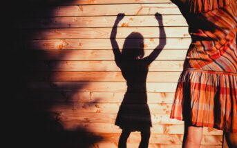 A person in a striped dress is partially visible on the right side, casting a shadow on a wooden wall. The shadow shows the person with arms raised, creating a playful, dynamic scene under warm lighting.