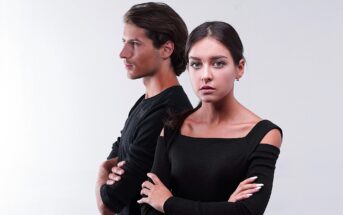 A man and woman stand back to back with arms crossed, both wearing black clothing. The woman looks directly at the camera with a neutral expression, while the man gazes off to the side. The background is plain white.