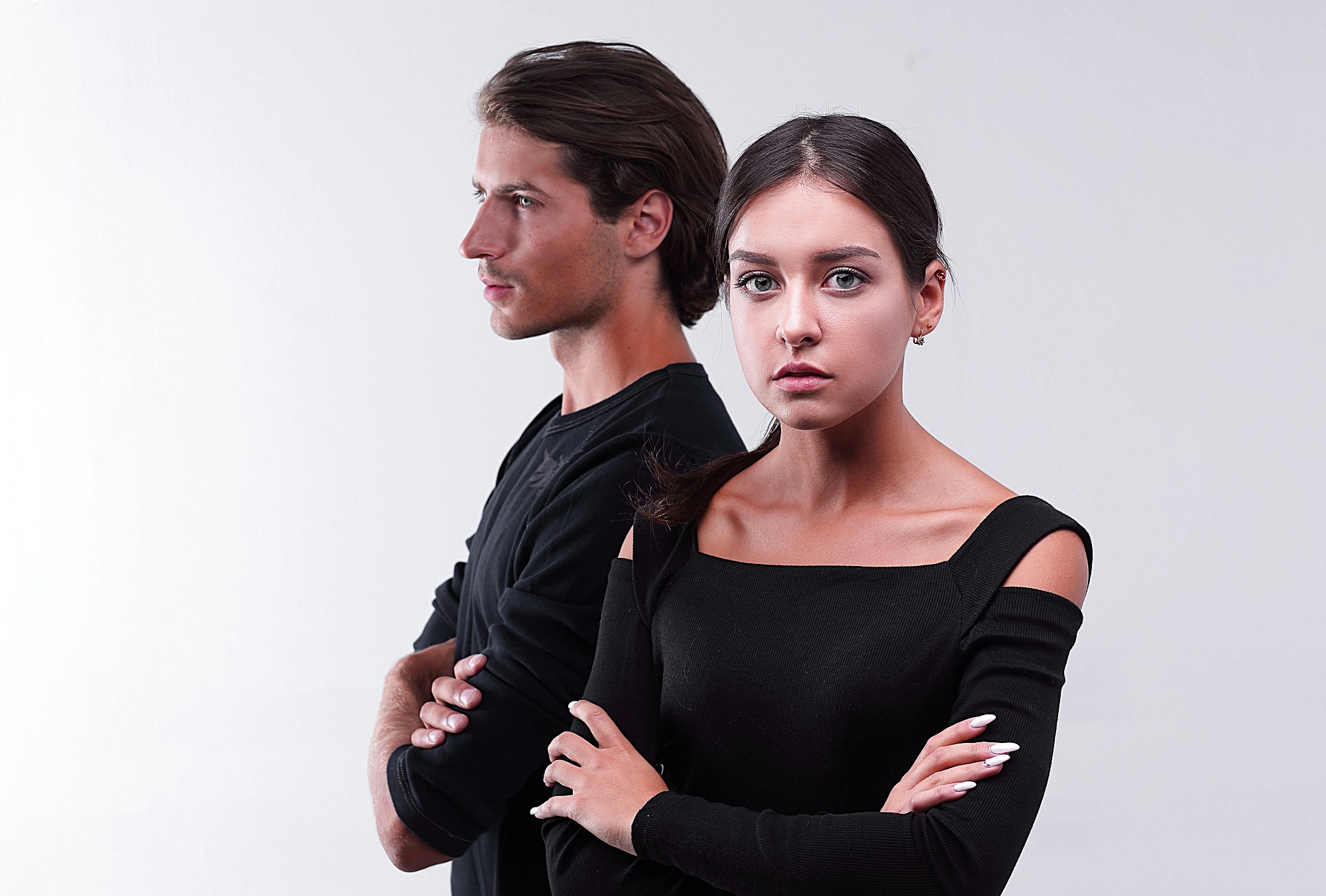 A man and woman stand back to back with arms crossed, both wearing black clothing. The woman looks directly at the camera with a neutral expression, while the man gazes off to the side. The background is plain white.