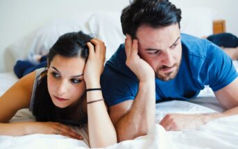 A man and woman lying on a bed, both looking thoughtful and concerned. They rest their heads in their hands, gazing towards the edge of the bed. The room has a neutral color scheme.