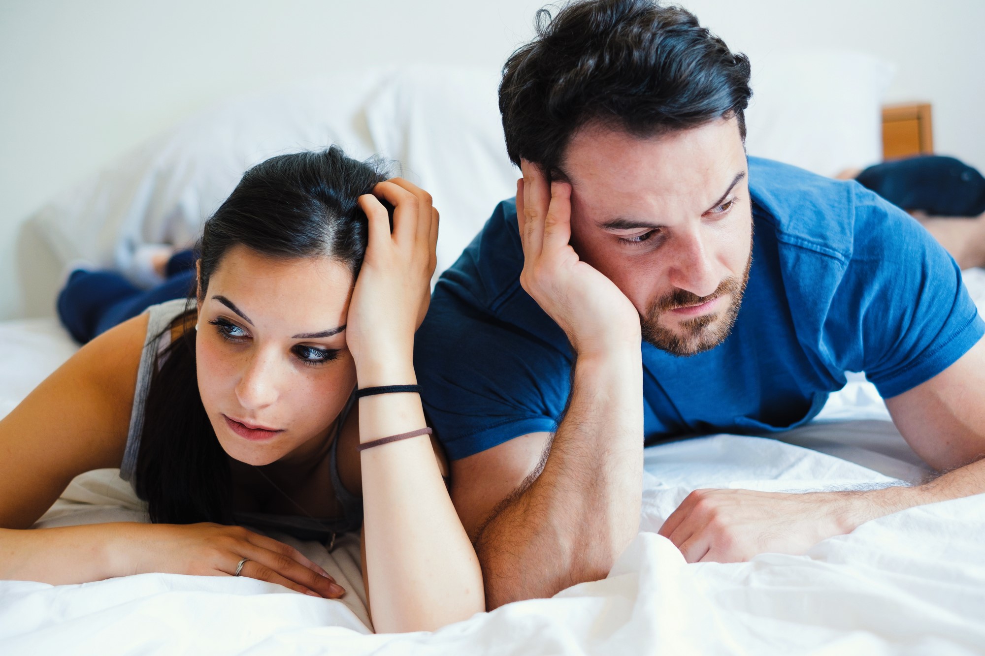 A man and woman lying on a bed, both looking thoughtful and concerned. They rest their heads in their hands, gazing towards the edge of the bed. The room has a neutral color scheme.