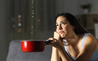 A woman sits on a couch indoors, holding a red pot to catch water dripping from above. She rests her chin on her hand, looking up with a frustrated expression. The room is dimly lit, with blurred lights visible in the background.