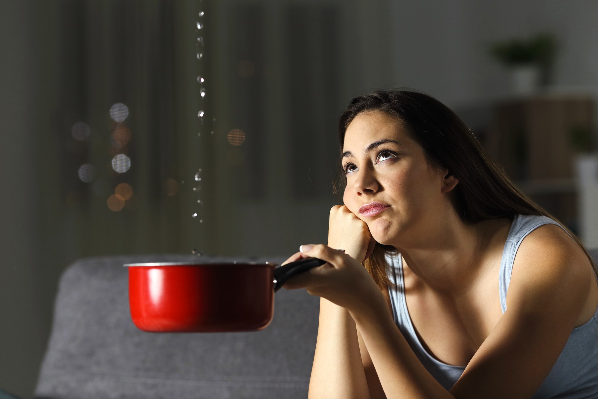 A woman sits on a couch indoors, holding a red pot to catch water dripping from above. She rests her chin on her hand, looking up with a frustrated expression. The room is dimly lit, with blurred lights visible in the background.