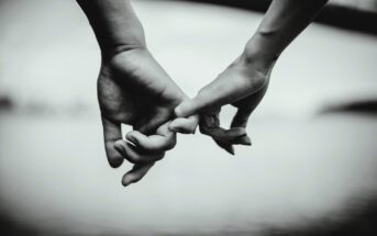 A black and white image of two hands gently hooking pinky fingers against a blurred background, symbolizing connection or promise.