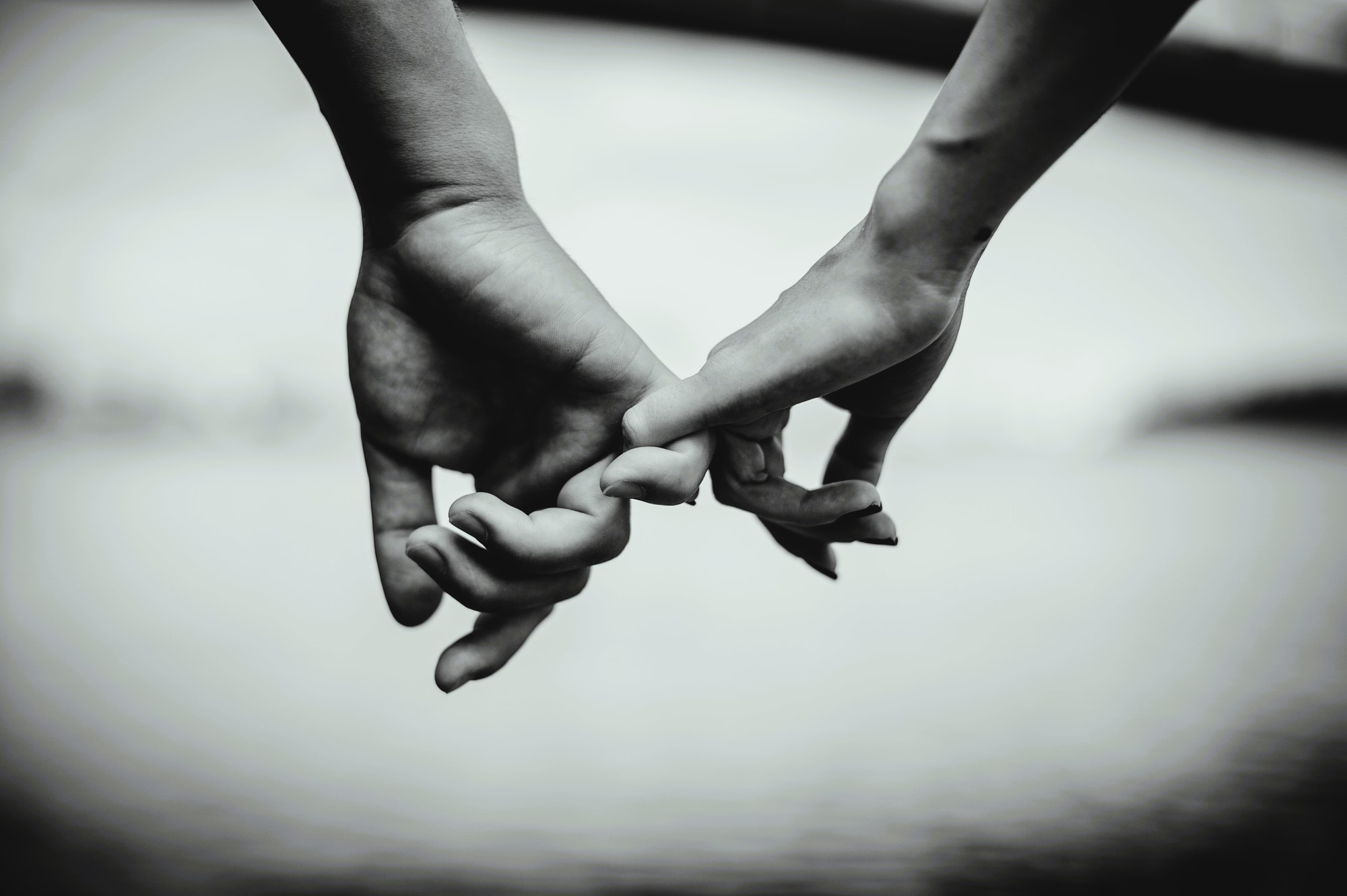 A black and white image of two hands gently hooking pinky fingers against a blurred background, symbolizing connection or promise.