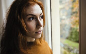 A woman with long red hair and a brown sweater gazes thoughtfully out a window. The background shows a soft focus of autumn trees with orange leaves, creating a serene and introspective mood.