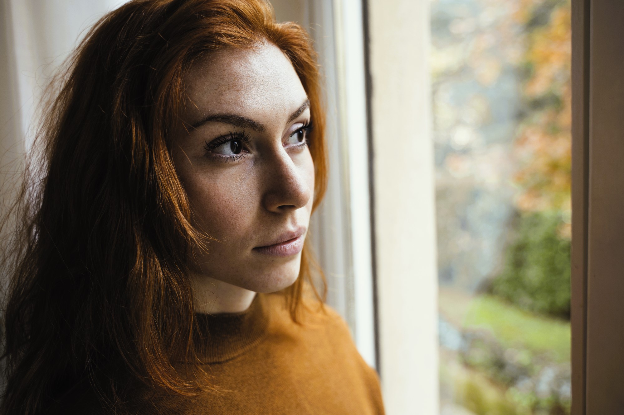 A woman with long red hair and a brown sweater gazes thoughtfully out a window. The background shows a soft focus of autumn trees with orange leaves, creating a serene and introspective mood.