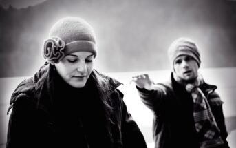 A black-and-white photo of a woman and a man outdoors. The woman, wearing a knit hat with a flower, looks down. The man, in a beanie and scarf, stands behind her with an arm extended. They appear to be in a mountainous area.